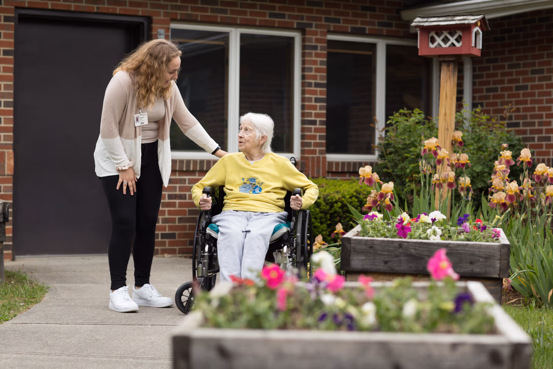 A caregiver with a name badge bends down to talk to an elderly woman in a wheelchair outside near flower beds and a brick building.