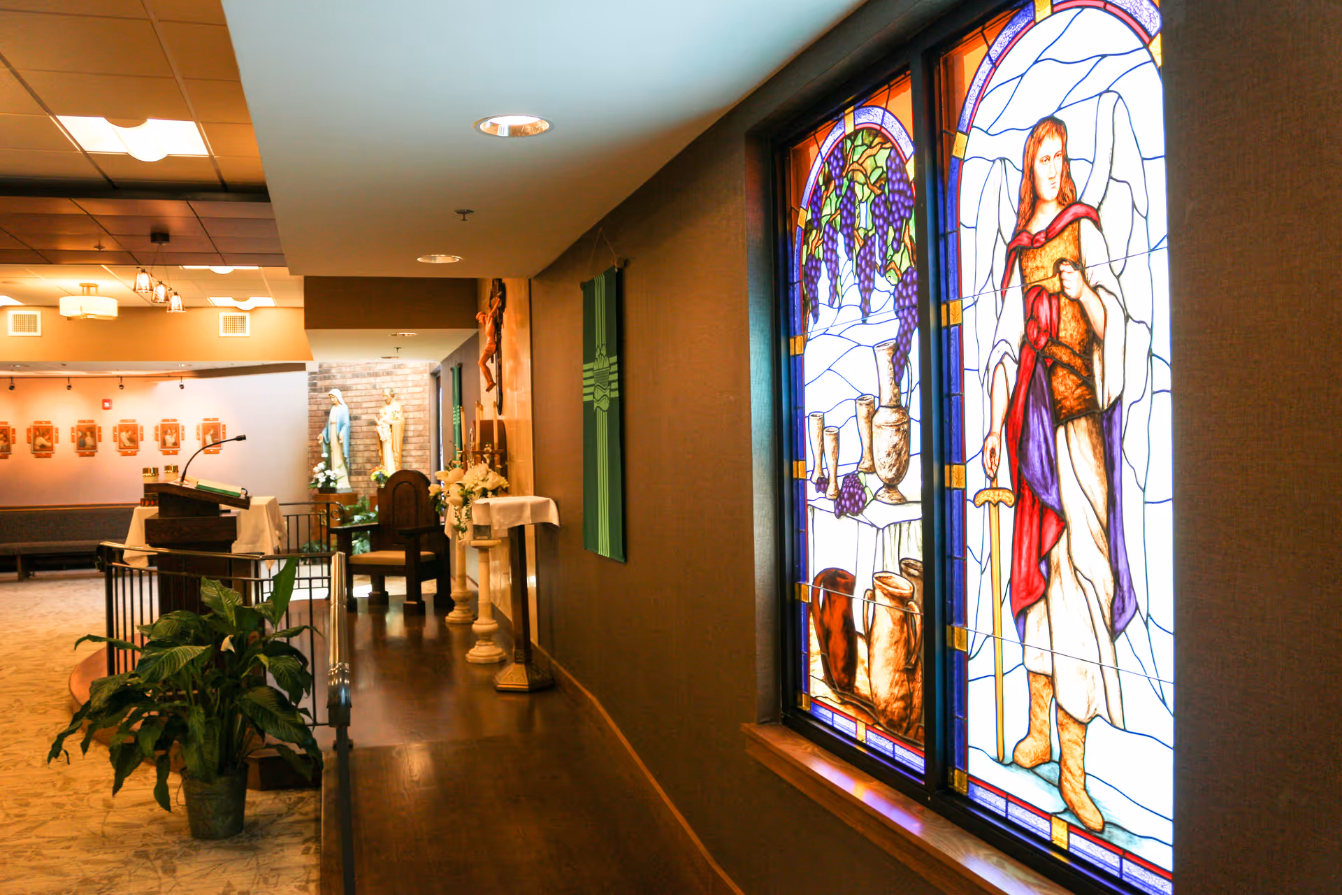 Interior view of a chapel or prayer room with stained glass windows depicting religious imagery, a lectern with a microphone, religious statues, and a plant near the entrance.