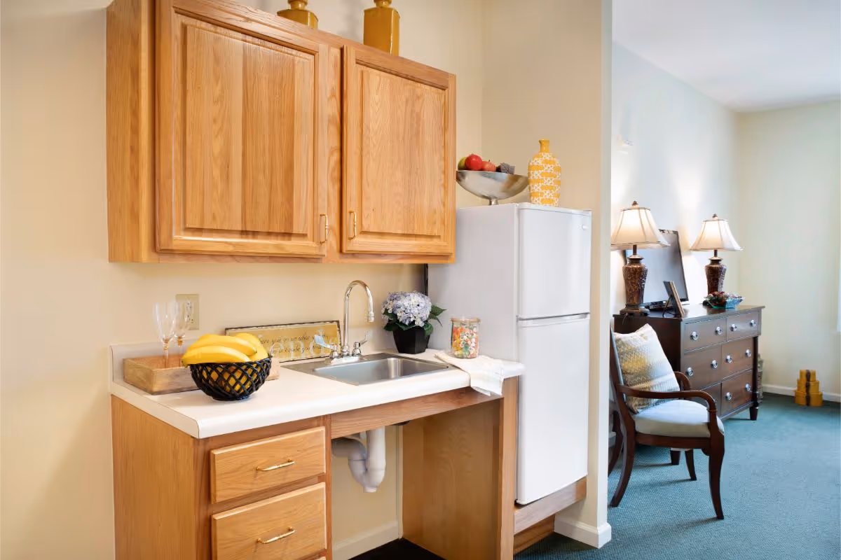 A small kitchenette area with wooden cabinets, a white countertop with a sink, a black fruit basket with bananas, two wine glasses, a decorative bowl with fruit on top of a white refrigerator, and a small potted plant. In the background, there is a wooden dresser with two table lamps and a cushioned armchair with a pillow in a carpeted room.