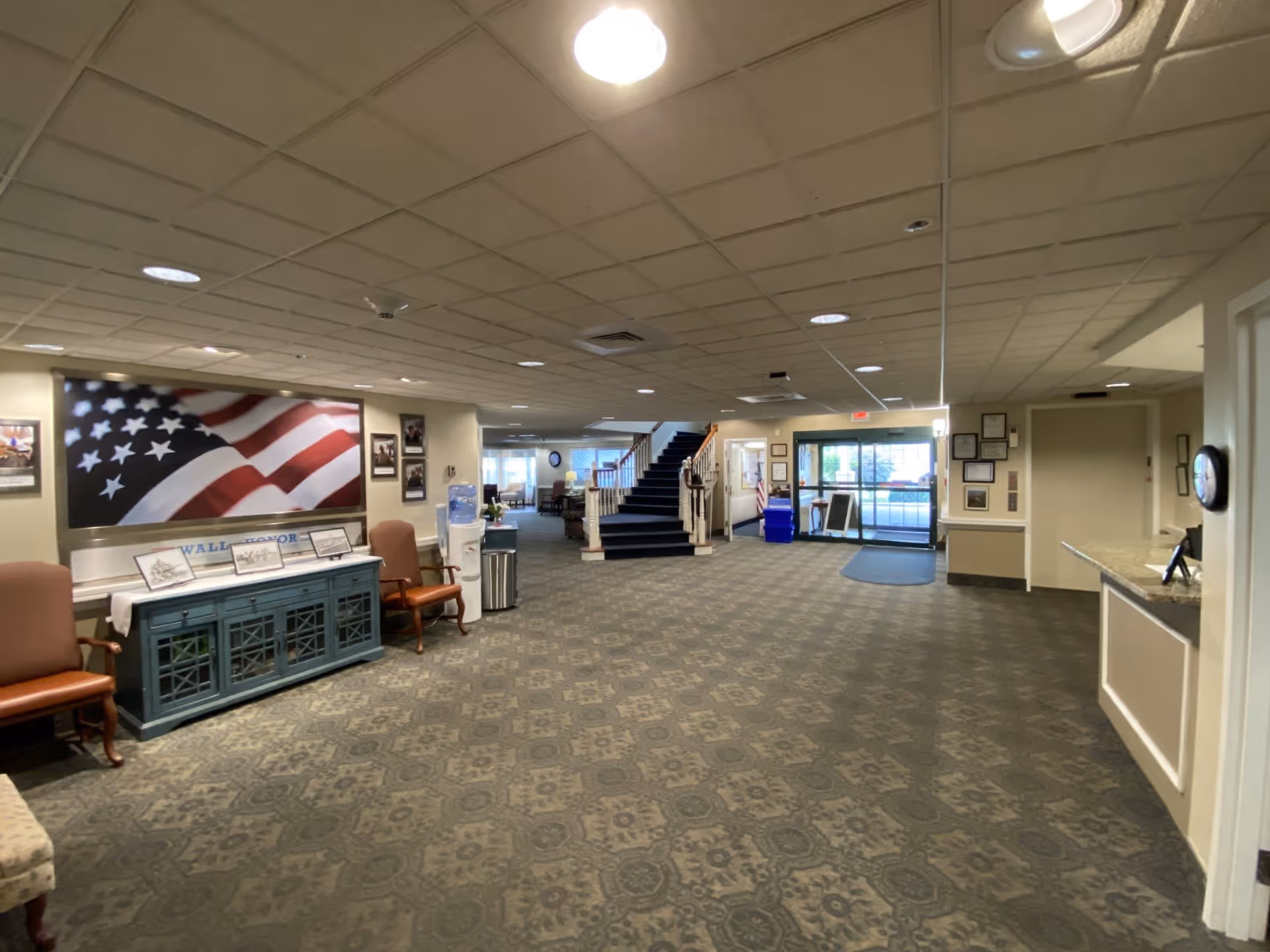 Spacious lobby area of Willowbrook Place featuring patterned carpet, a large American flag wall art, chairs, a water cooler, a staircase leading to an upper floor, and a glass entrance door with natural light coming through.
