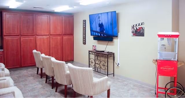 A small theater room with six beige upholstered chairs arranged in two rows facing a wall-mounted flat screen TV. There is a red popcorn machine on the right side and a decorative metal table beneath the TV. The walls are light-colored and there is a large wooden cabinet on the left side of the room.