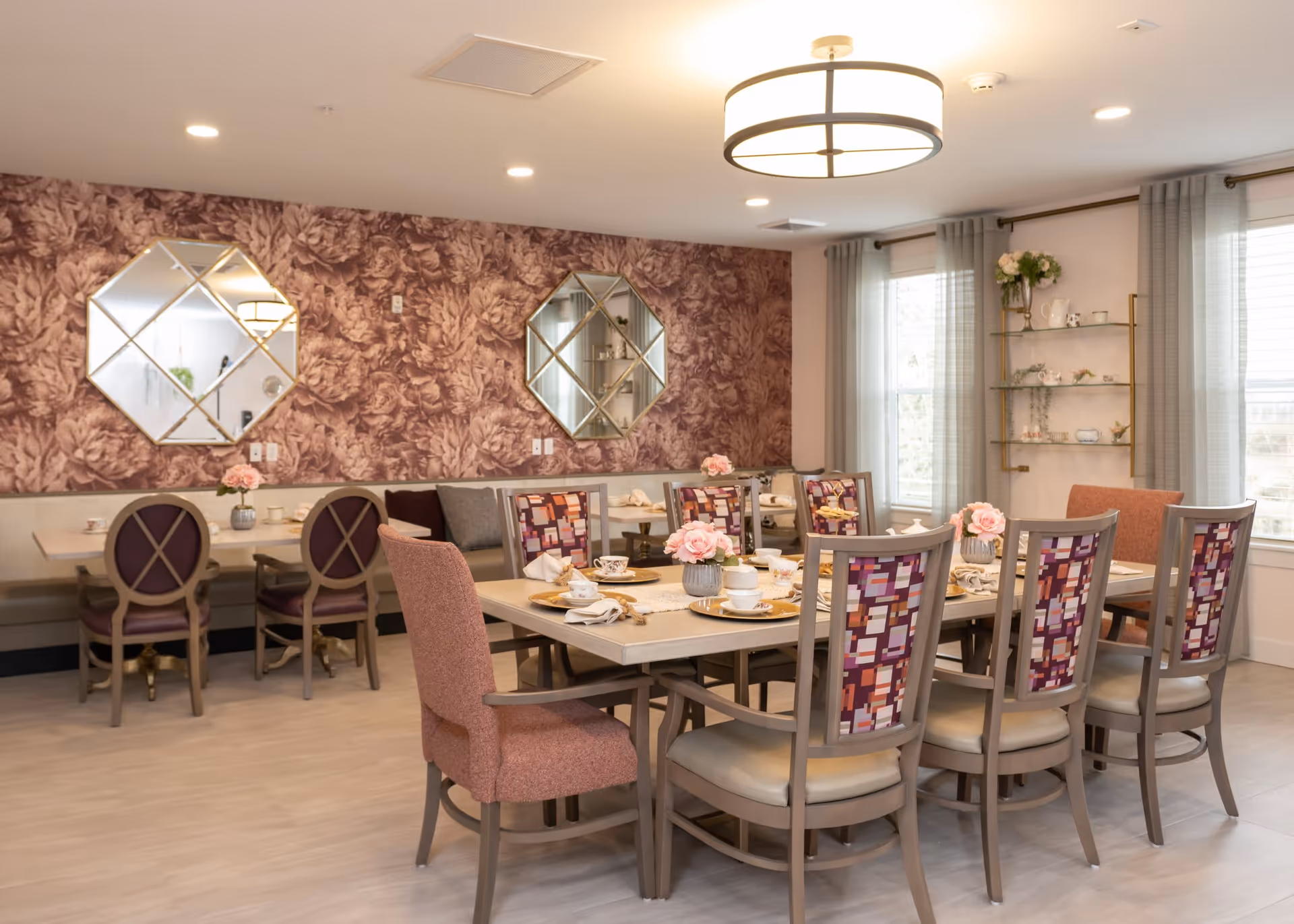 A well-lit dining room with a large rectangular table set for a meal, featuring pink floral centerpieces and patterned chairs. The room has floral wallpaper on one wall with two decorative mirrors, light gray curtains on windows, and a modern ceiling light fixture.