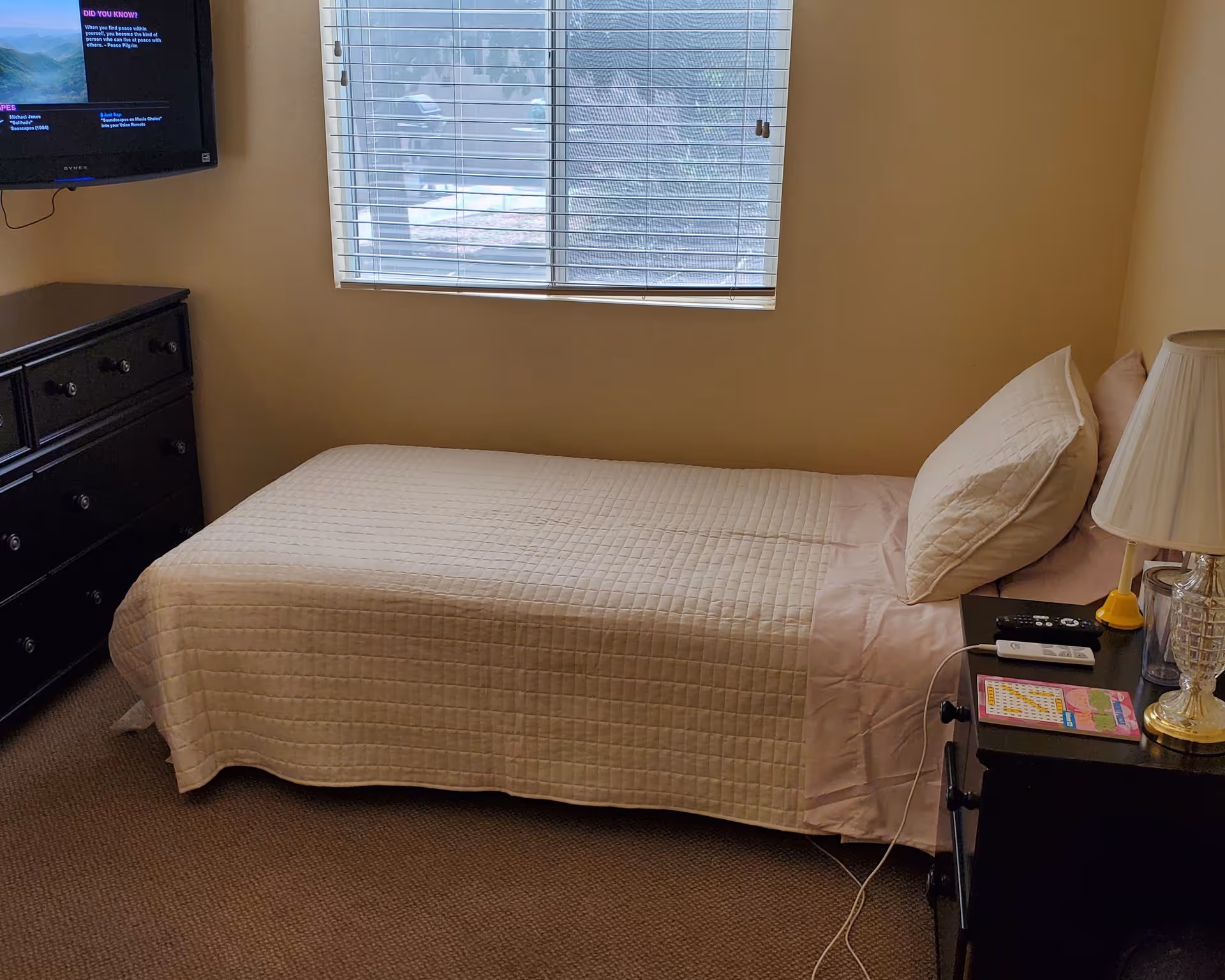 A small bedroom with a single bed covered in a light beige quilt and pink sheets. There is a window with blinds above the bed. To the right of the bed is a black nightstand with a lamp, a remote control, a cup, and a puzzle book. To the left of the bed is a black dresser with multiple drawers. A wall-mounted TV is visible in the upper left corner of the image.