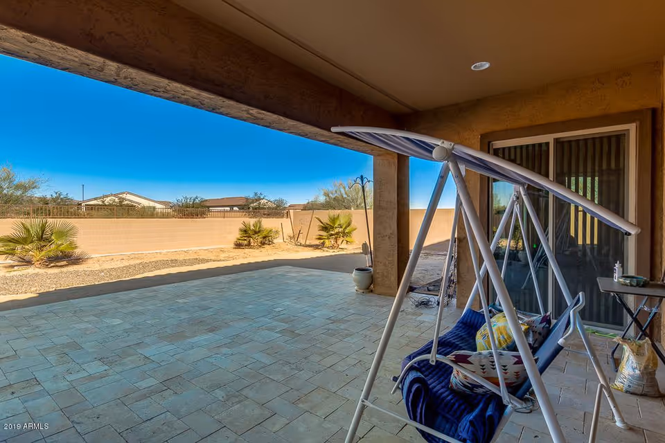 Covered outdoor patio area with a white metal swing chair that has a blue cushion and colorful pillows. The patio has tiled flooring and overlooks a fenced backyard with desert landscaping including small palm plants and gravel. The sky is clear and blue.
