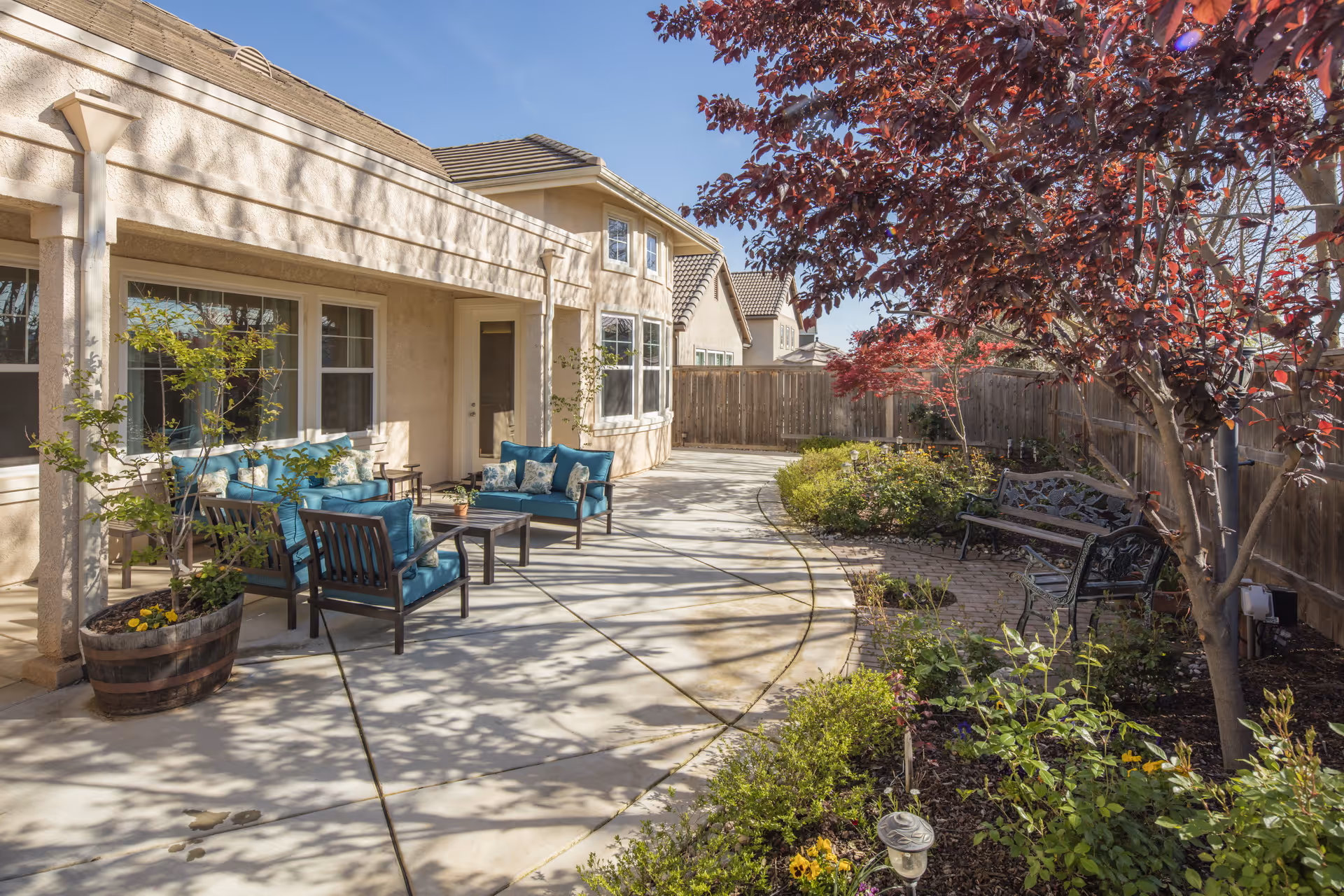 Outdoor patio area of a residential care facility with cushioned seating, a coffee table, potted plants, a curved concrete walkway, garden beds with various plants and flowers, a wooden fence, and a tree with red leaves under a clear blue sky.