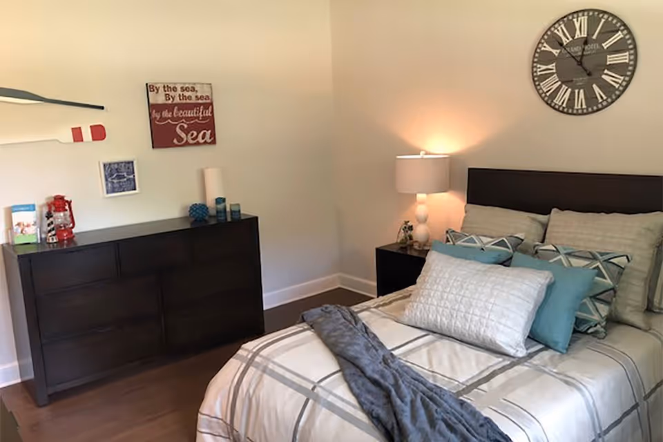 A neatly furnished bedroom with a made bed, pillows, bedside lamp and a dresser under coastal-themed wall decor.