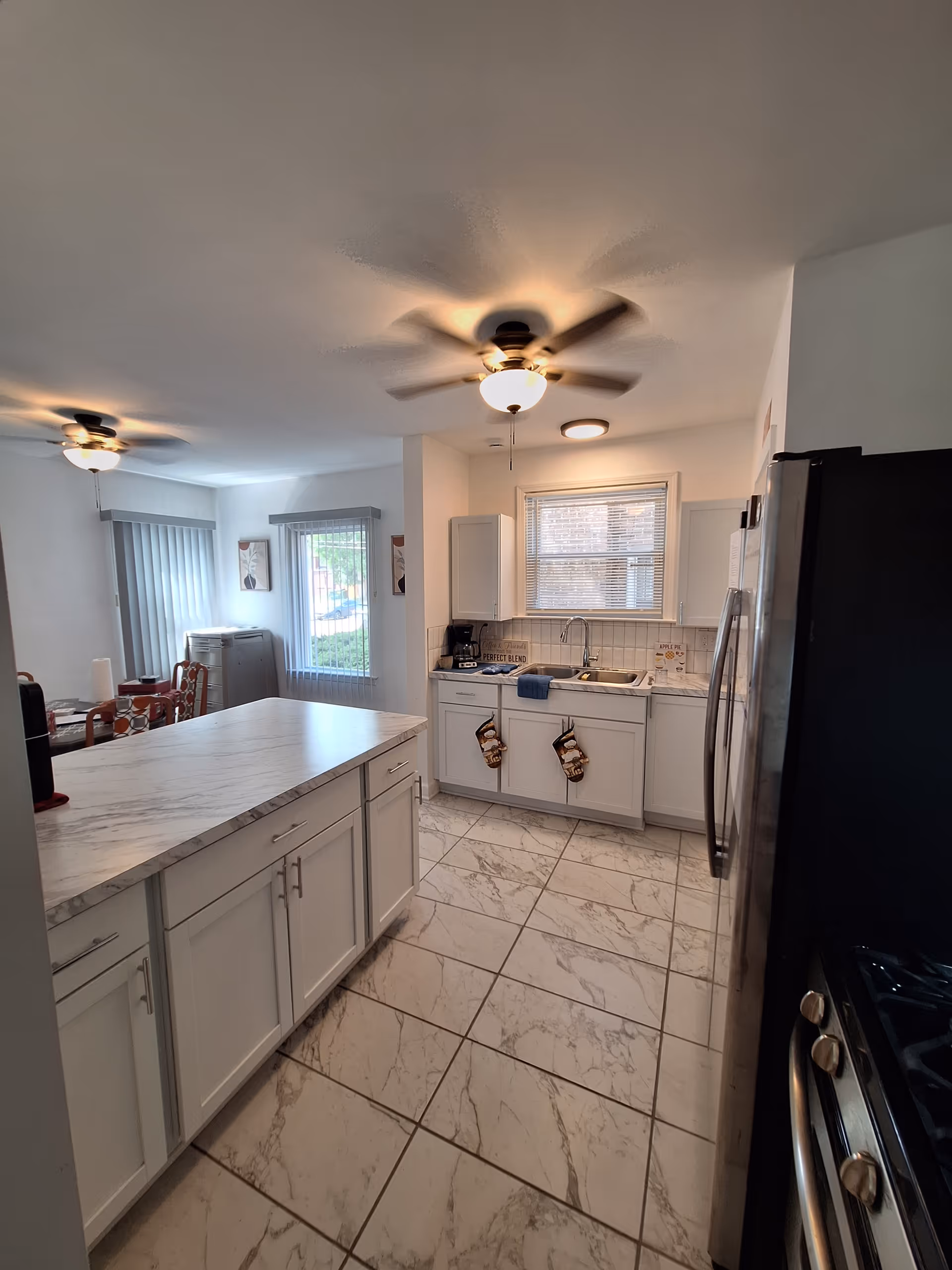 Bright modern kitchen with a large marble-look island, white cabinets, tiled floor, stainless appliances and ceiling fans.