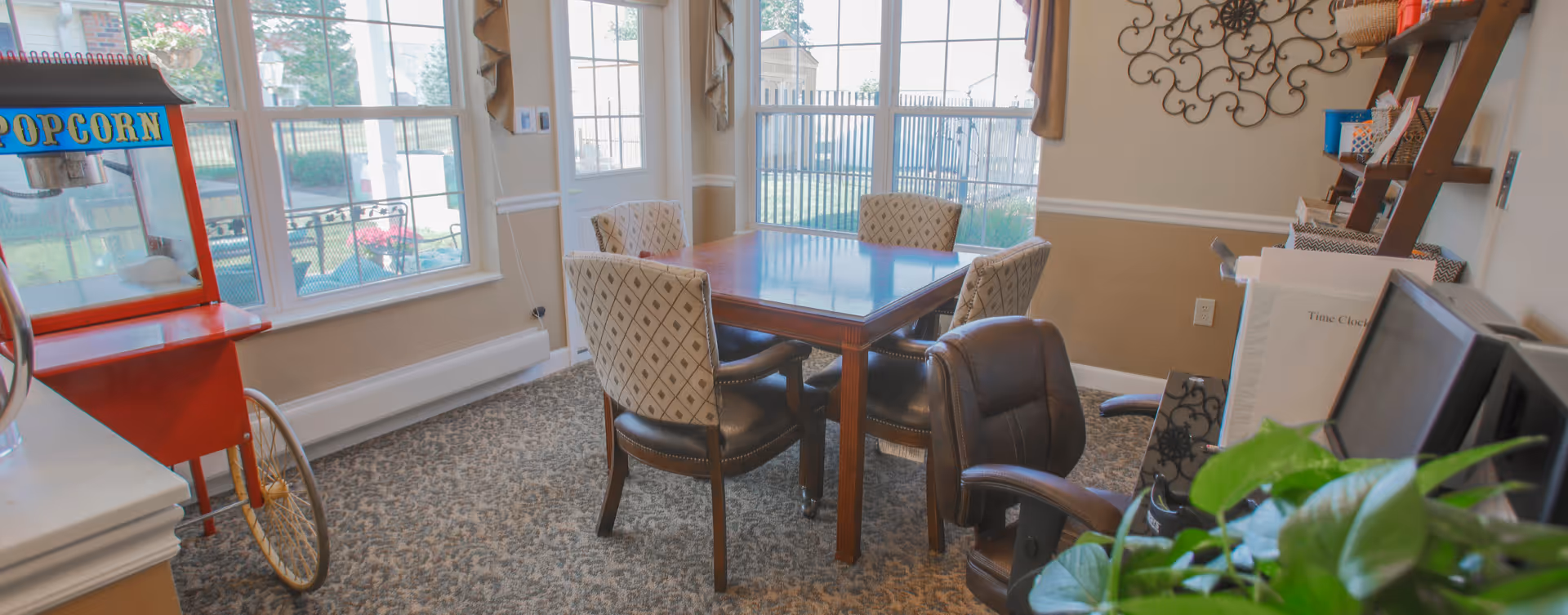 A cozy interior room with a wooden table surrounded by four upholstered chairs with diamond patterns. There is a brown leather office chair in the foreground. Large windows and a glass door let in natural light, showing a fenced outdoor area. A red popcorn machine is visible on the left side, and a decorative wall piece and shelving unit with baskets and papers are on the right wall. A green plant is partially visible in the bottom right corner.