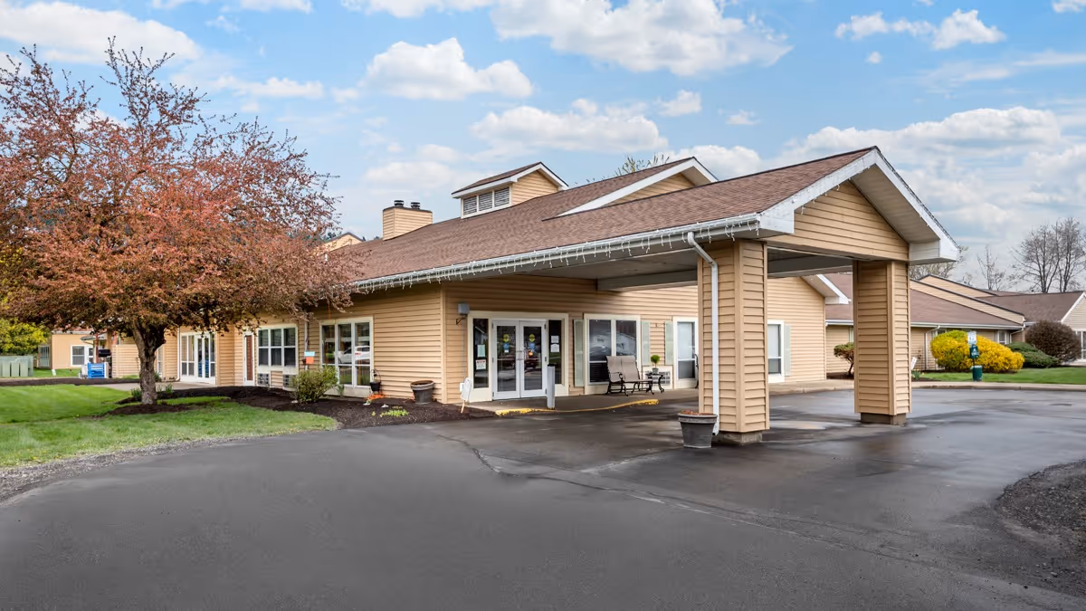 Entrance of a single-story beige senior living facility with a covered porte-cochère, paved driveway, and landscaped lawn.