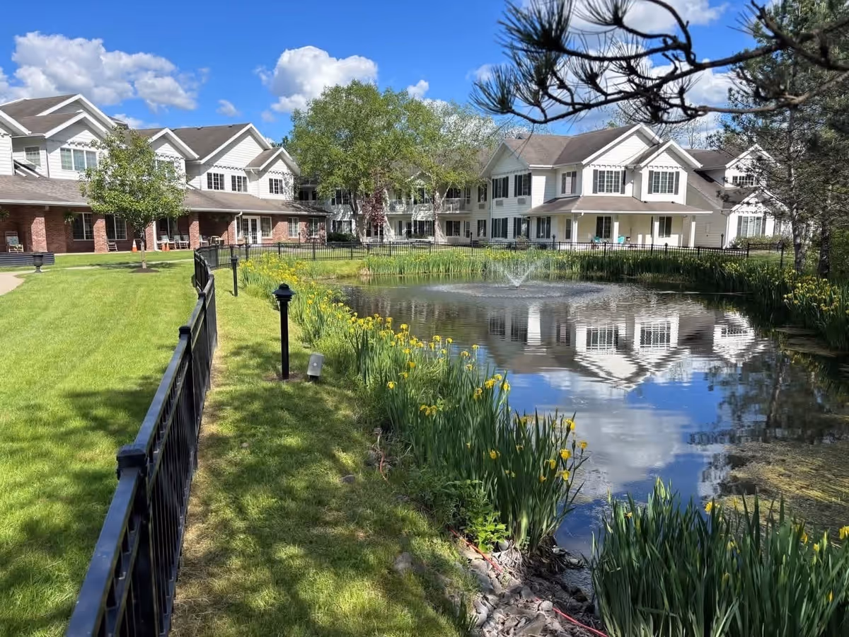 A serene outdoor scene at Holiday Maple Downs featuring a pond with a fountain in the center, surrounded by green grass and yellow flowers. There is a black metal fence along the edge of the pond, and large white residential buildings with multiple windows and porches are visible in the background under a blue sky with scattered clouds.