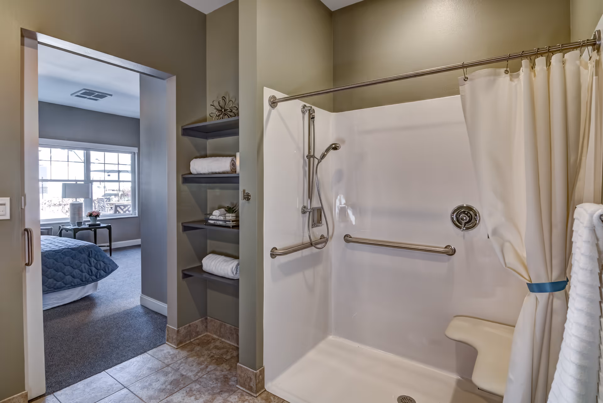 Bathroom with a walk-in shower featuring a fold-down seat, grab bars, and a handheld showerhead. There are shelves with rolled towels and decorative items next to the shower. Through the doorway, a bedroom with a large window, a bed with a blue quilt, and a side table with a lamp and flowers is visible.