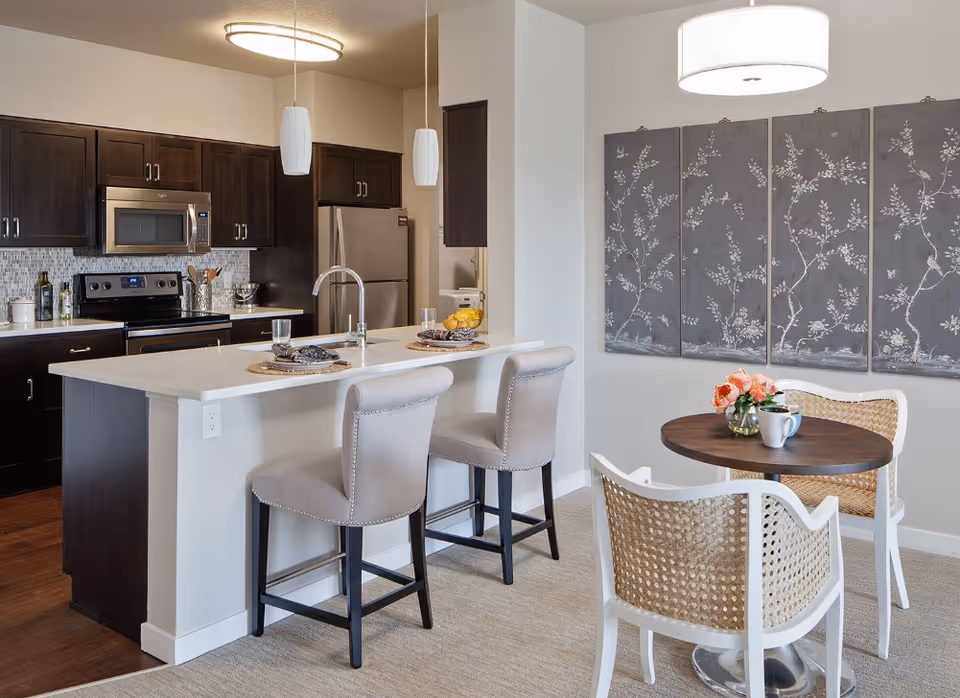Open kitchen with a white island, two upholstered bar stools, dark cabinets, stainless appliances, and a small round dining table with two chairs.