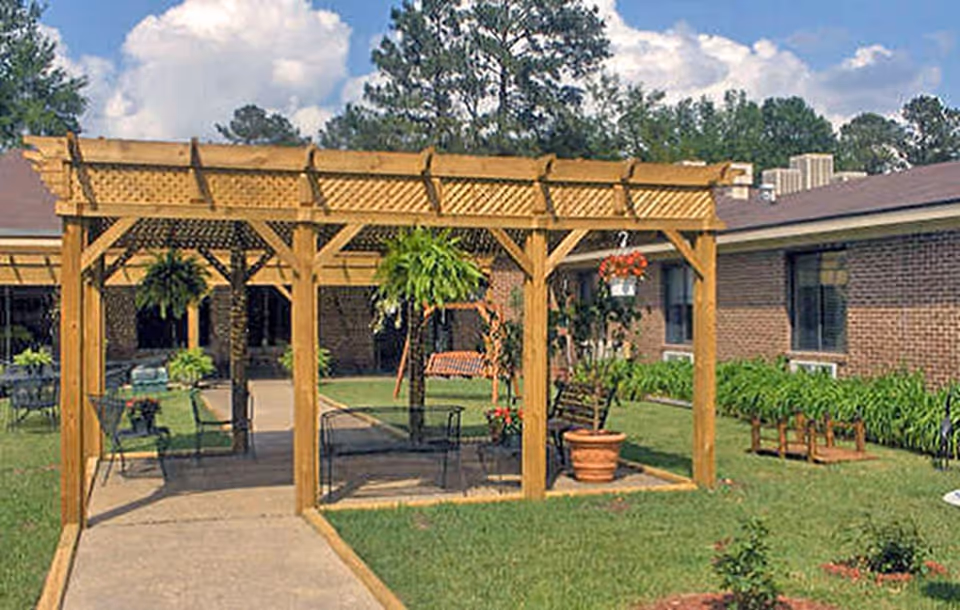 Outdoor courtyard area with a wooden pergola structure, hanging plants, potted flowers, metal chairs, and a swing bench. The area is surrounded by a brick building and green grass with some shrubs and trees in the background under a partly cloudy sky.