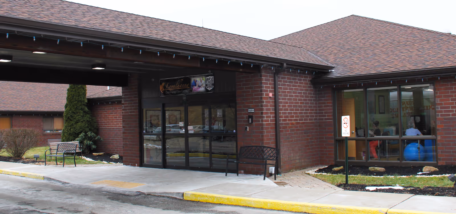 Exterior view of Loyalhanna Care Center showing the main entrance with glass doors, brick walls, a covered drop-off area, benches, and a no smoking sign near a window where people are visible inside.