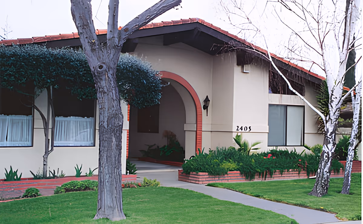 Front exterior of a single-story building with an arched entryway, lawn, trees, and the house number 2405 visible.