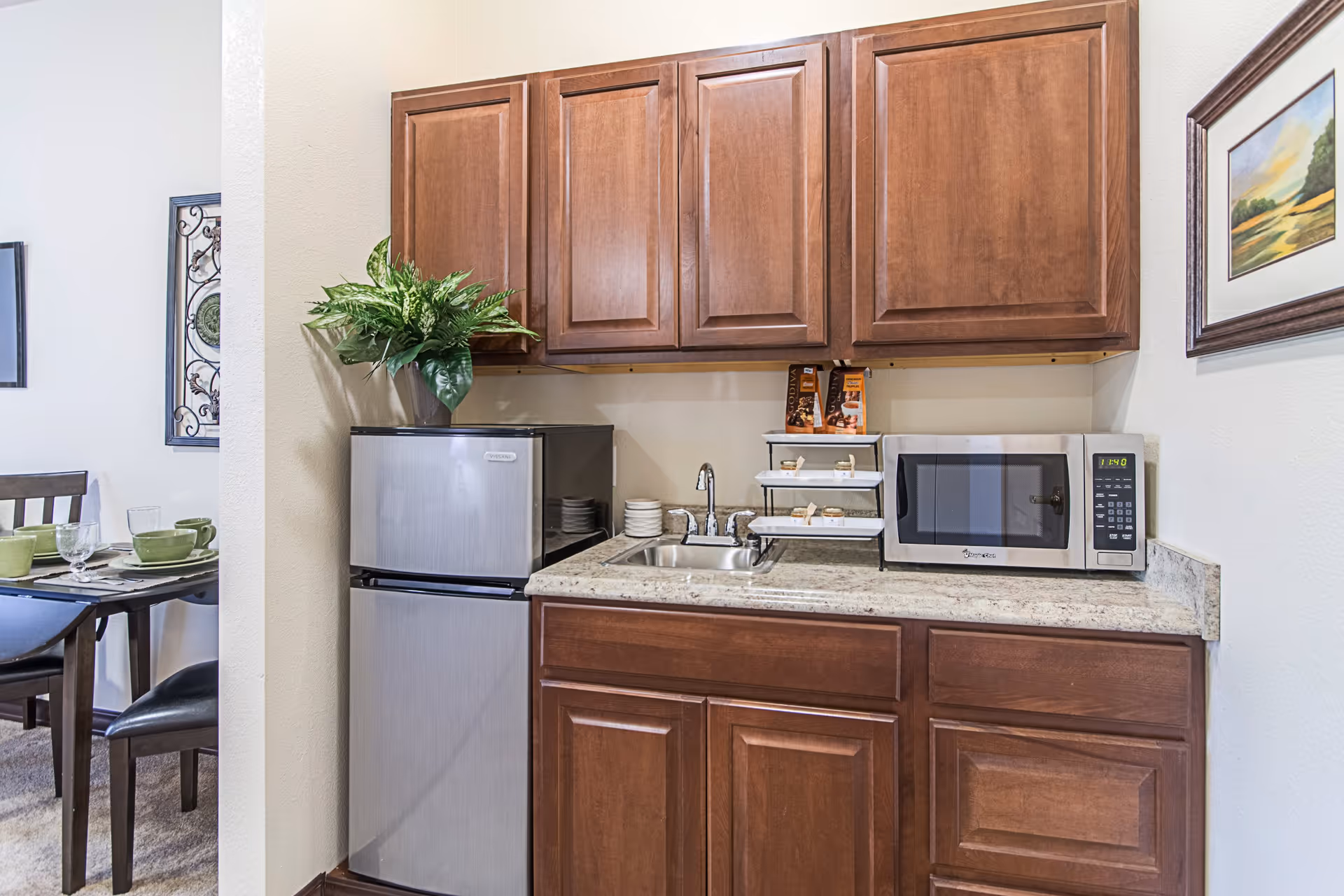 Small kitchen area with wooden cabinets, a stainless steel mini refrigerator, a microwave, a small sink, and a countertop with a tiered tray holding snacks. A dining table with green dishes and chairs is partially visible to the left. A framed landscape painting hangs on the wall to the right.