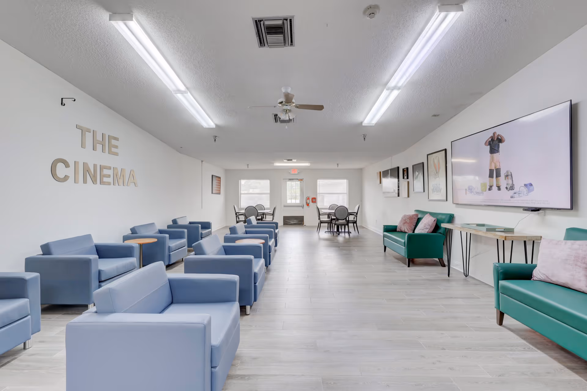 A spacious, well-lit common area with multiple blue armchairs and small wooden tables arranged in rows on the left side. On the right side, there are green sofas with pink cushions and a large flat-screen TV mounted on the wall displaying an image of a person. The wall on the left has the words 'THE CINEMA' in large letters. At the far end, there are two round tables with chairs near windows letting in natural light. The ceiling has fluorescent lights and a ceiling fan.