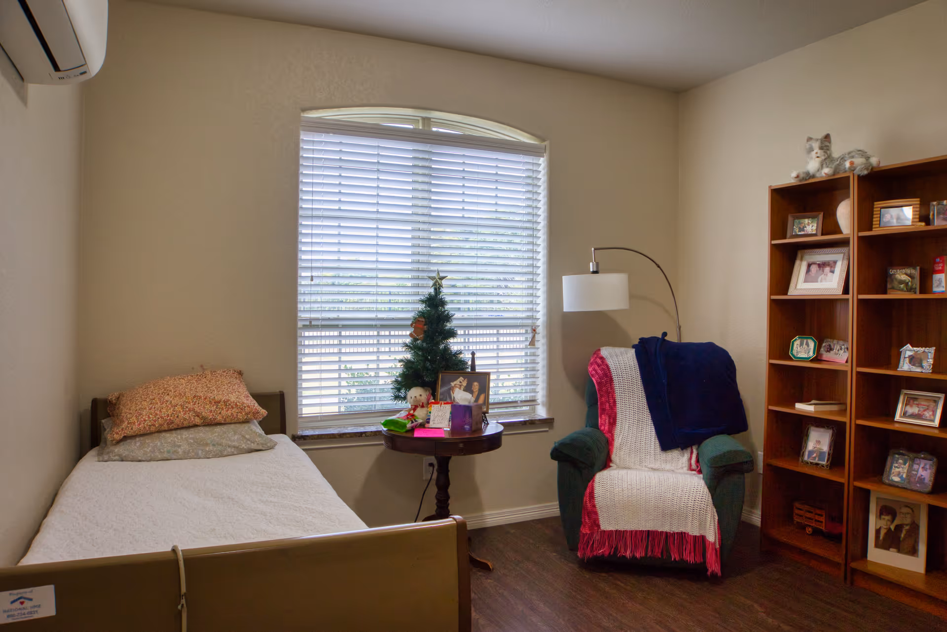 A small bedroom with a single bed on the left side, a window with blinds in the center, and a green armchair with a white and pink throw blanket and a dark blue blanket draped over it on the right. A round wooden side table with a small Christmas tree, framed photos, and other small items is placed in front of the window. A wooden bookshelf filled with framed photos and decorative items is on the right wall. The room has wooden flooring and beige walls.