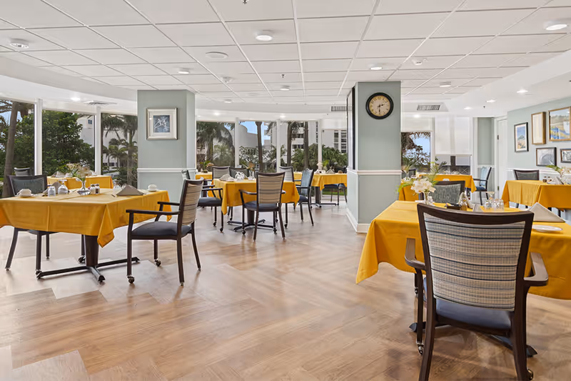 A bright dining room with multiple tables covered in yellow tablecloths, each set with plates, cups, and silverware. The room has large windows allowing natural light and views of palm trees and buildings outside. The floor is wooden, and there are several chairs around each table. A clock is mounted on a pillar in the center of the room, and framed artwork decorates the walls.