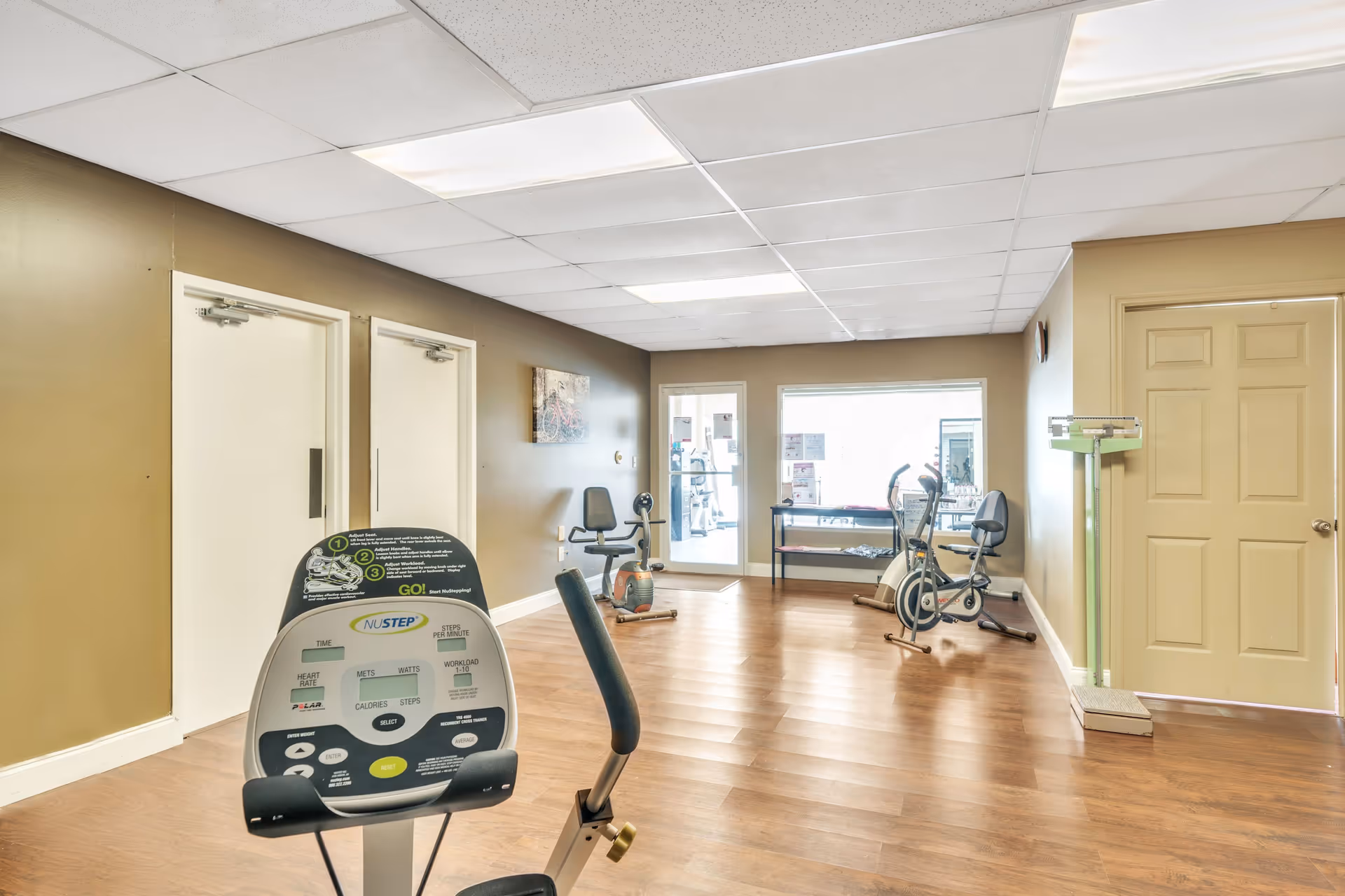 A small exercise room with light brown walls and wood flooring. The room contains exercise equipment including a NuStep recumbent cross trainer, a stationary bike, and another exercise bike. There is a large window and a glass door letting in natural light. A green scale is positioned near a closed beige door on the right side of the room.