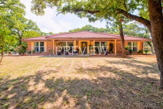 Single-story brick building with a red tile roof surrounded by large trees and a dry grassy yard under a clear sky.