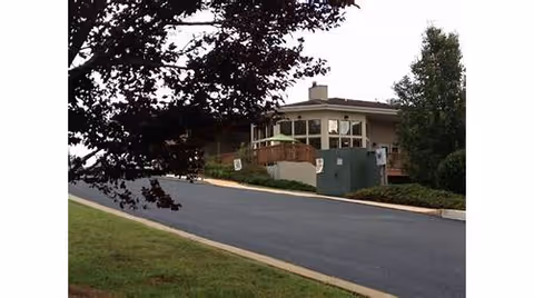 View of a single-story building with a sloped driveway leading up to it, surrounded by trees and greenery.