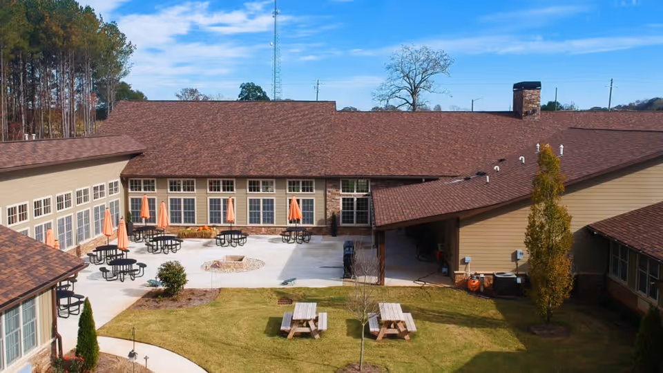 Outdoor courtyard area of The Retreat at Canton featuring multiple round tables with orange umbrellas, a fire pit in the center, picnic tables on a grassy area, and surrounding buildings with brown roofs and large windows under a partly cloudy sky.