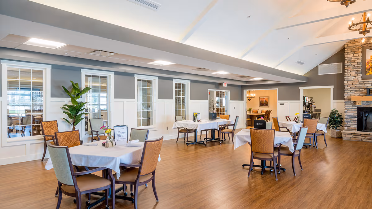 A spacious dining room with several tables covered in white tablecloths, each surrounded by chairs. The room has wood flooring, gray and white walls with large windows, and a stone fireplace on the right side. There are decorative plants and framed artwork on the walls, creating a warm and inviting atmosphere.