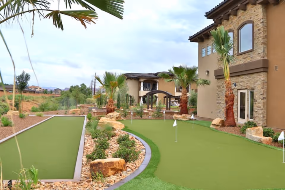 Outdoor putting green area with small flags and palm trees, surrounded by landscaped rocks and plants, adjacent to a building with stone and stucco exterior walls under a cloudy sky.