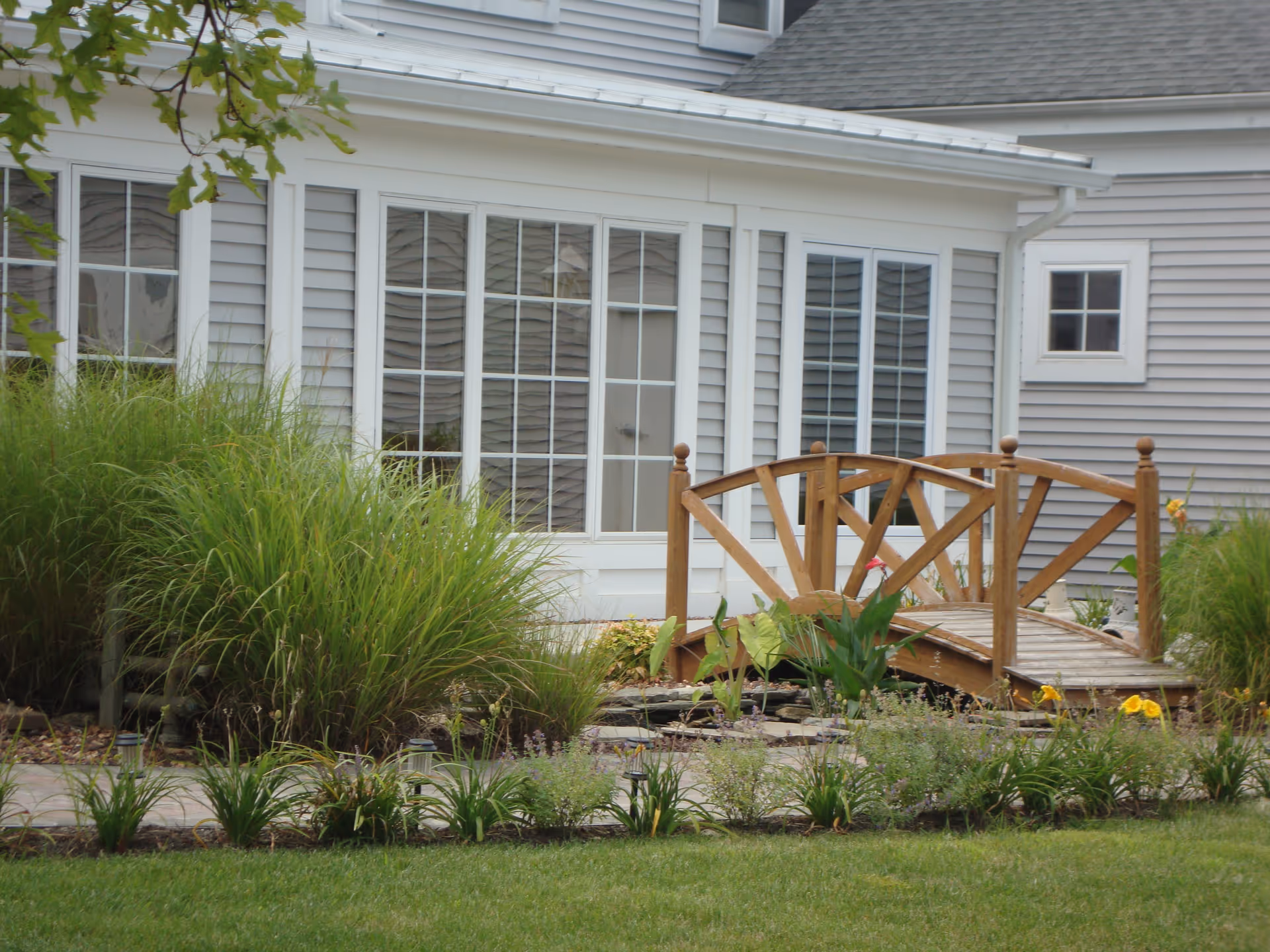 A small wooden footbridge over a garden pond in front of a building with large windows and gray siding. The garden features green grass, various plants, and flowers.