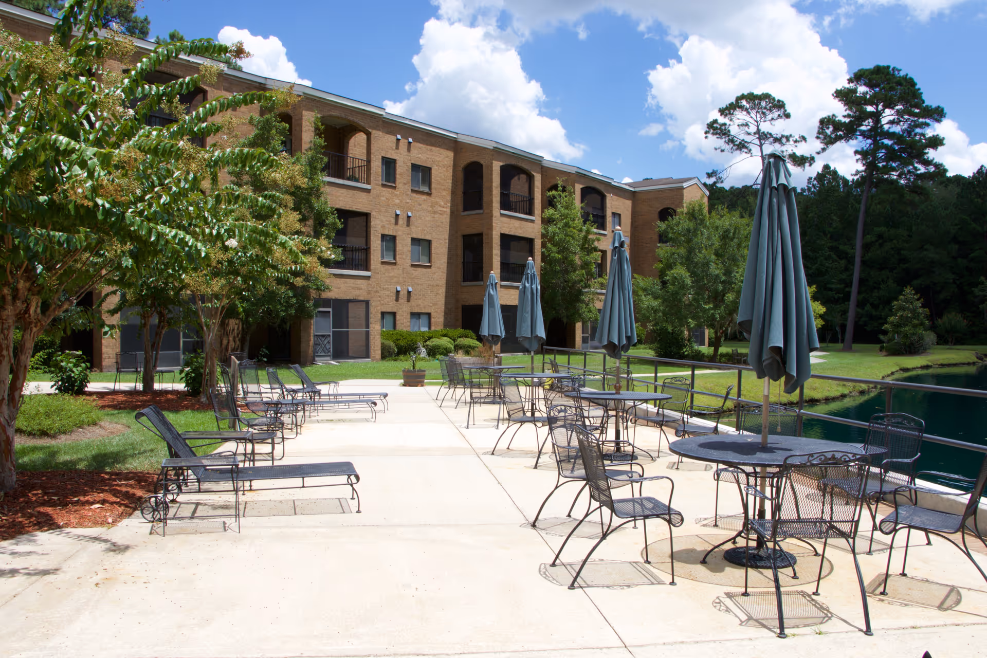 Outdoor patio area with metal tables and chairs, each table shaded by a closed umbrella. The patio is adjacent to a three-story brick building with balconies, surrounded by trees and greenery under a partly cloudy sky.