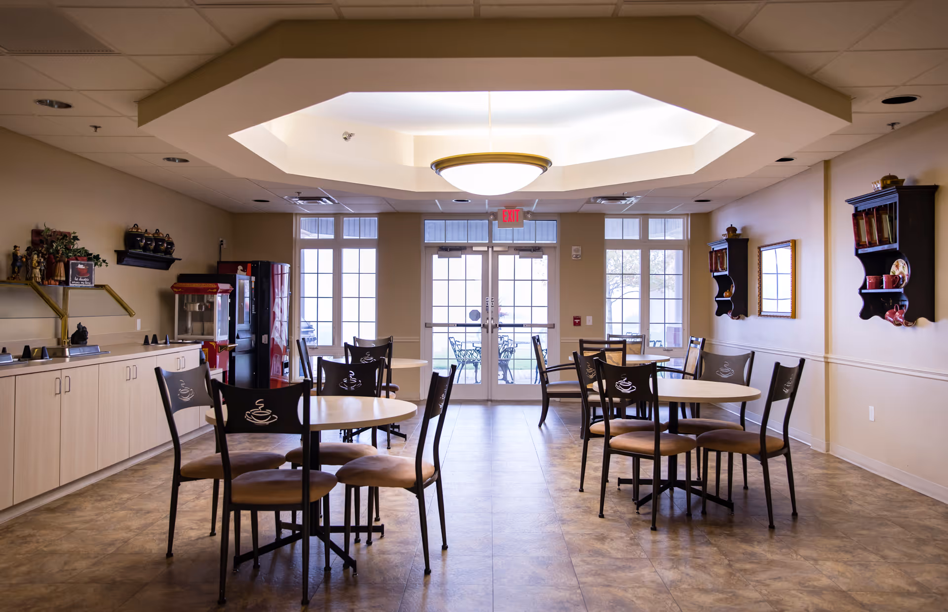 A bright dining area in a senior living facility with round tables and chairs featuring coffee cup designs on the backrests. The room has a large ceiling light fixture, beige walls, and a tiled floor. There is a counter with cabinets on the left side, a popcorn machine, and a vending machine near the back wall with large windows and glass doors leading outside to a patio with metal chairs and tables.
