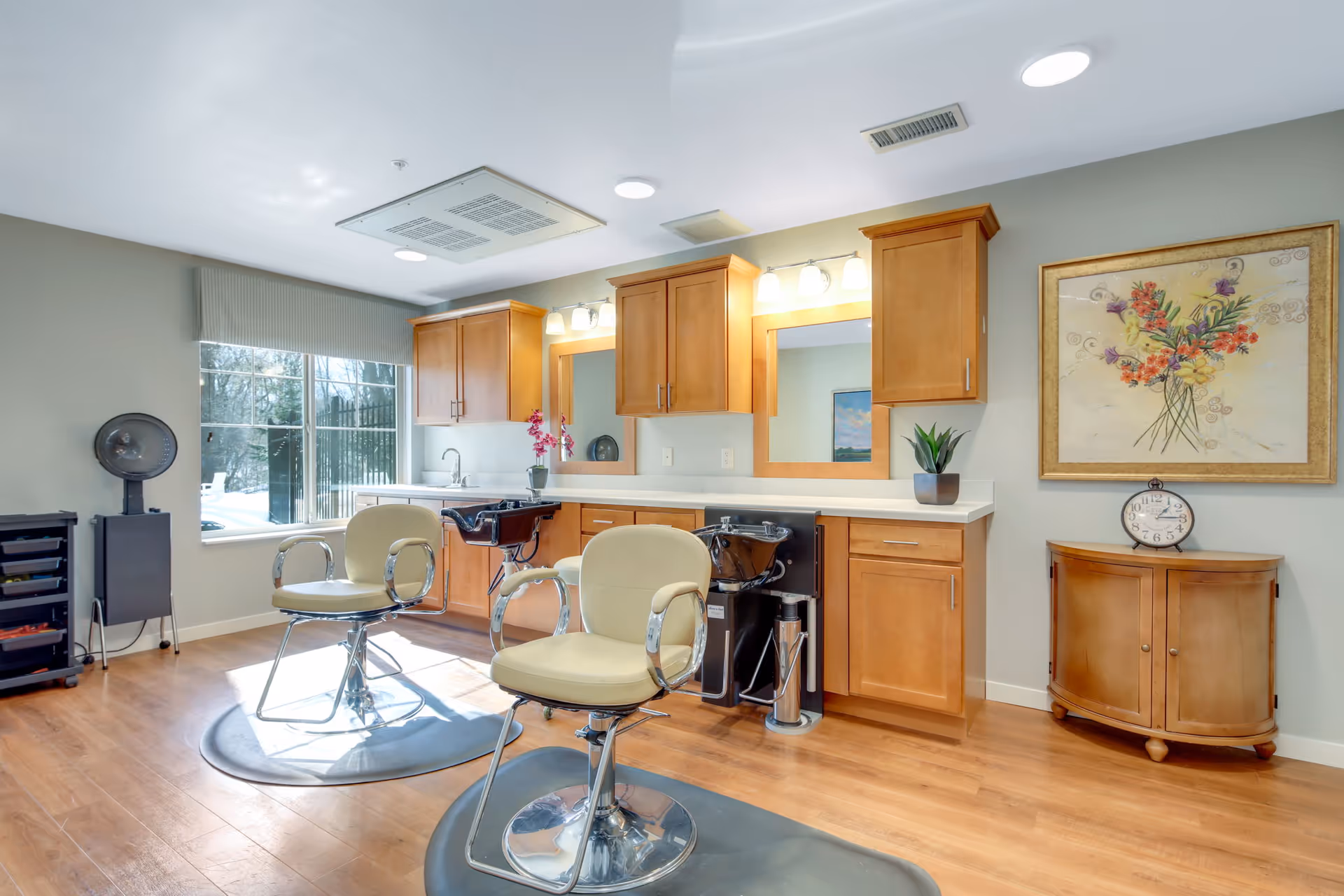 Interior view of a hair salon area in a senior living facility with two salon chairs, two hair washing sinks, wooden cabinets, a large window, a small wooden cabinet with a clock and a framed floral painting on the wall.
