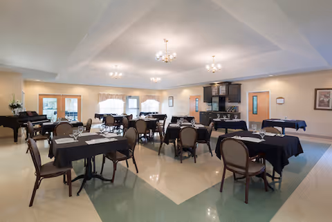 Spacious dining room with multiple tables dressed in black tablecloths and place settings, chairs, chandeliers, and a piano in the corner.