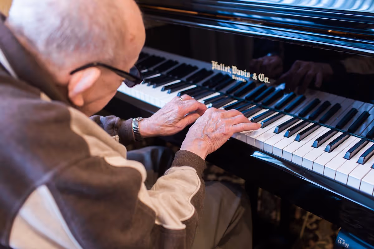 An elderly man wearing glasses and a brown jacket is playing a black Hallet Davis & Co. piano, focusing on his hands pressing the keys.