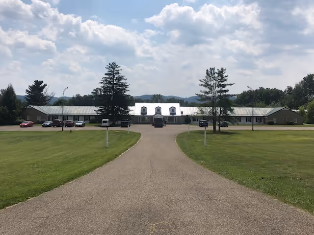 A long driveway leads to a single-story building with a metal roof and dormer windows, surrounded by green lawns and trees under a partly cloudy sky.