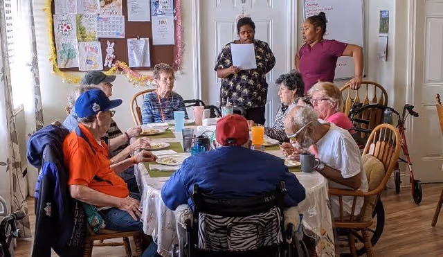 A group of elderly individuals seated around a dining table in a well-lit room, eating and drinking. Two caregivers stand nearby, one holding a piece of paper and the other observing. The room has wooden chairs, a bulletin board with various papers and decorations, and a walker is visible in the background.