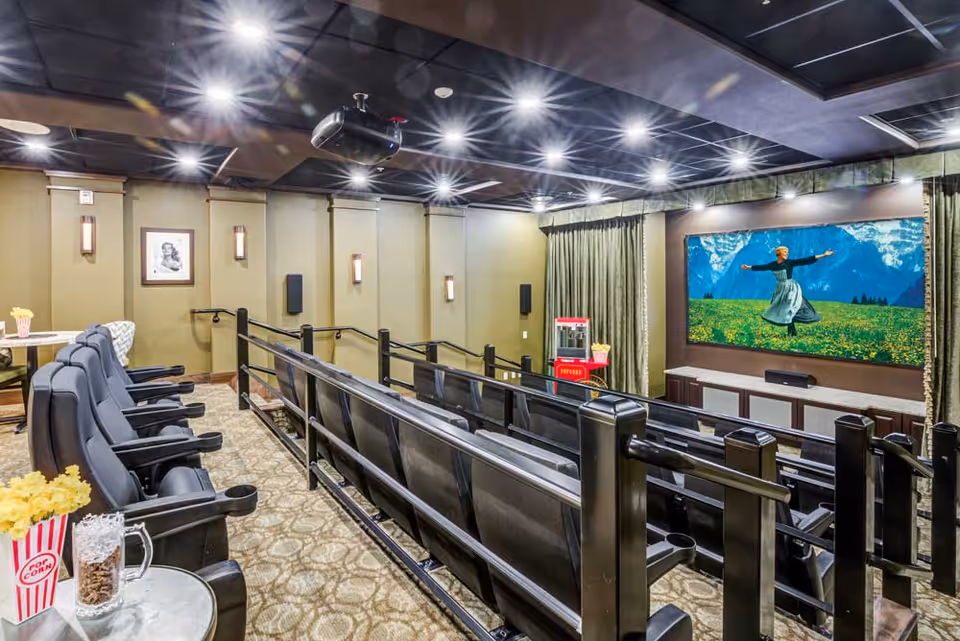 Cozy private movie theater with tiered reclining seats facing a large screen showing a woman in a field, plus a popcorn machine and snack table.