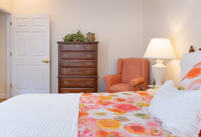 A cozy bedroom featuring a bed with a colorful floral bedspread and white pillows, a wooden chest of drawers with decorative items on top, a coral upholstered armchair, a white table lamp on a bedside table, and a white door slightly ajar against a light-colored wall.