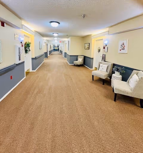 A long, well-lit hallway in a senior living facility with beige carpet and walls painted in cream and blue. Several doors line the hallway, some decorated with wreaths and signs. There are cushioned chairs and small tables with plants and decorative items along the walls. Ceiling lights illuminate the corridor.