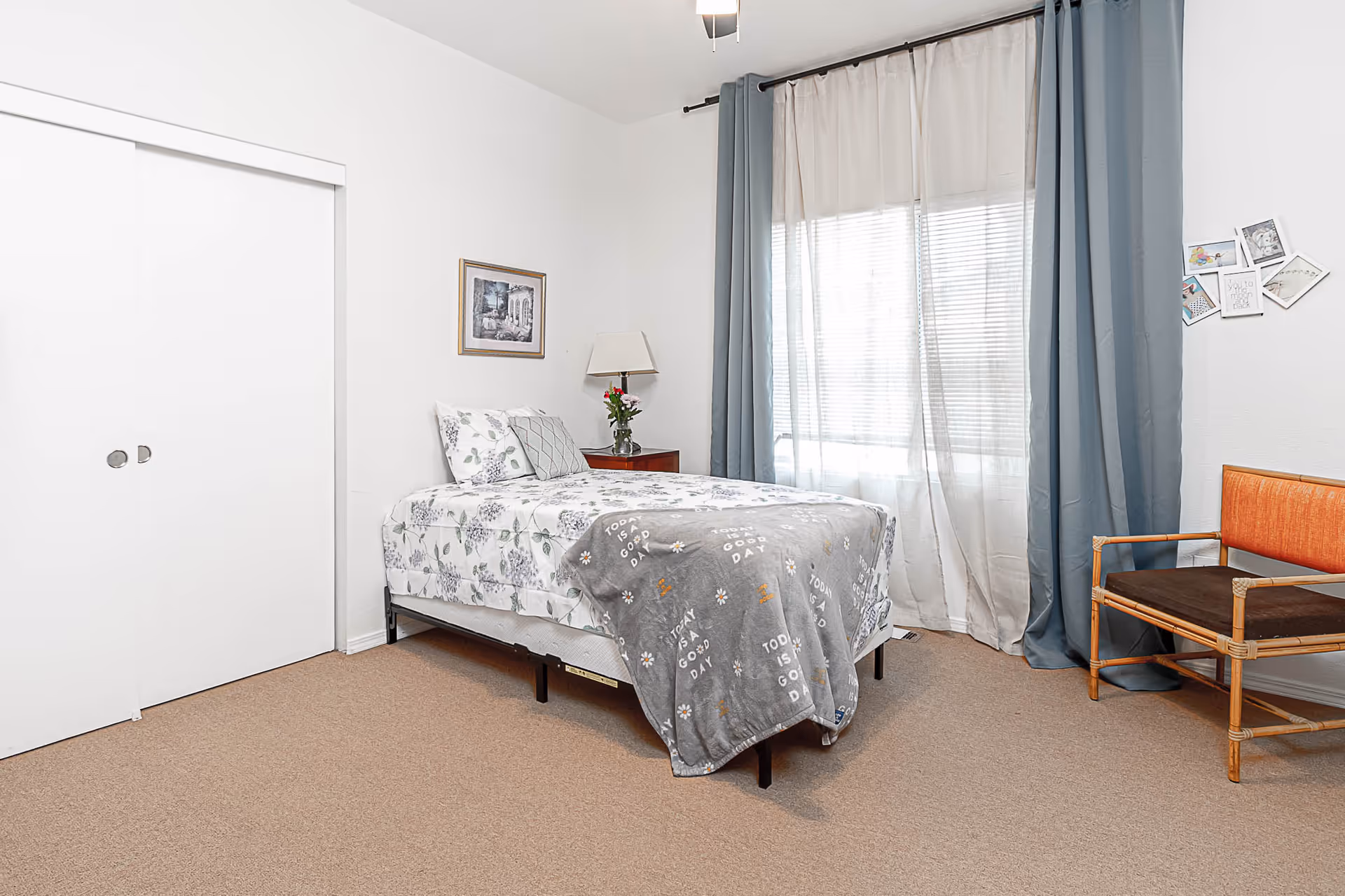 A simple bedroom with a single bed covered in floral bedding and a gray blanket that says 'Today is a good day.' There is a wooden nightstand with a lamp and a vase of flowers next to the bed. A framed picture hangs on the wall above the nightstand. The room has beige carpet, white walls, a large window with sheer white and blue curtains, and a wooden chair with orange and brown cushions. A white sliding closet door is visible on the left side of the room.