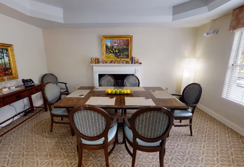 A dining room with a wooden table set for six, a bowl of apples centerpiece, upholstered chairs, and a decorative fireplace with artwork on the far wall.