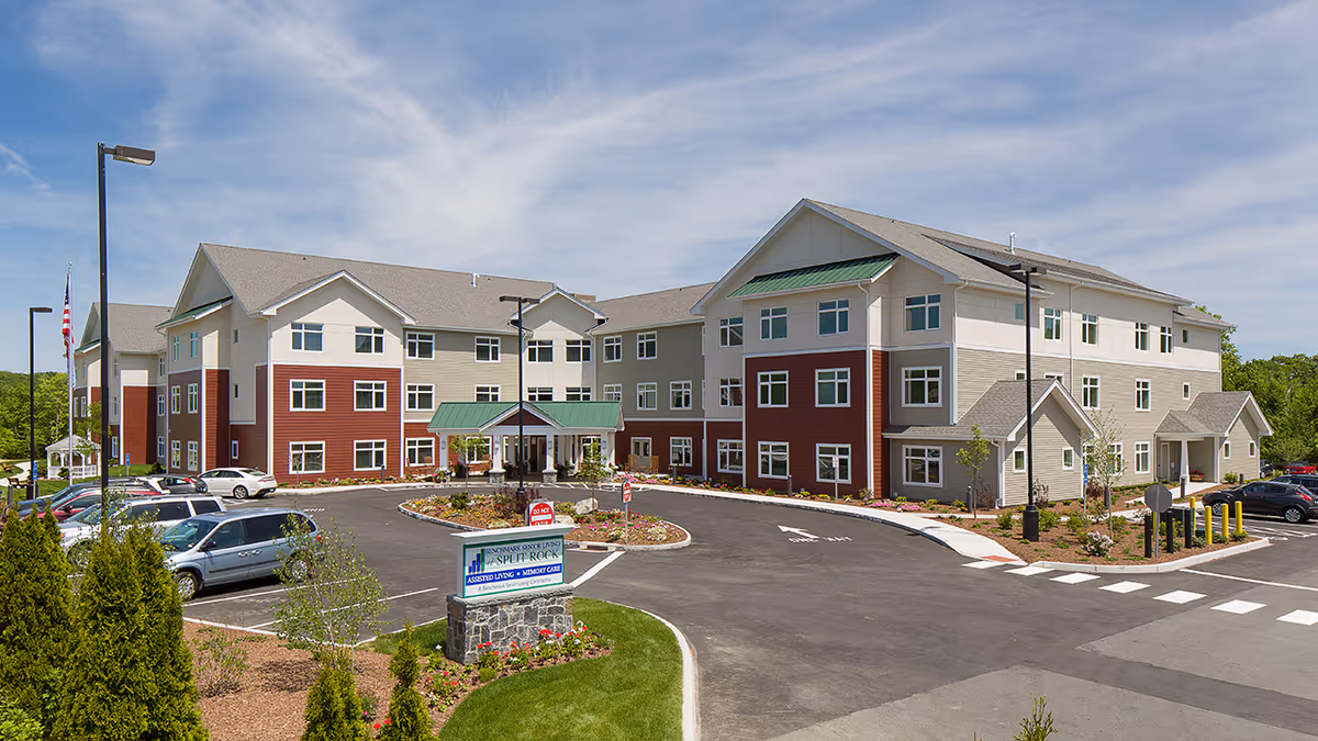 Exterior view of Benchmark Senior Living at Split Rock, a three-story building with beige, white, and red siding. The building has multiple windows, green awnings, and a covered entrance. There is a parking lot with several cars and landscaped areas with trees and shrubs under a partly cloudy sky.