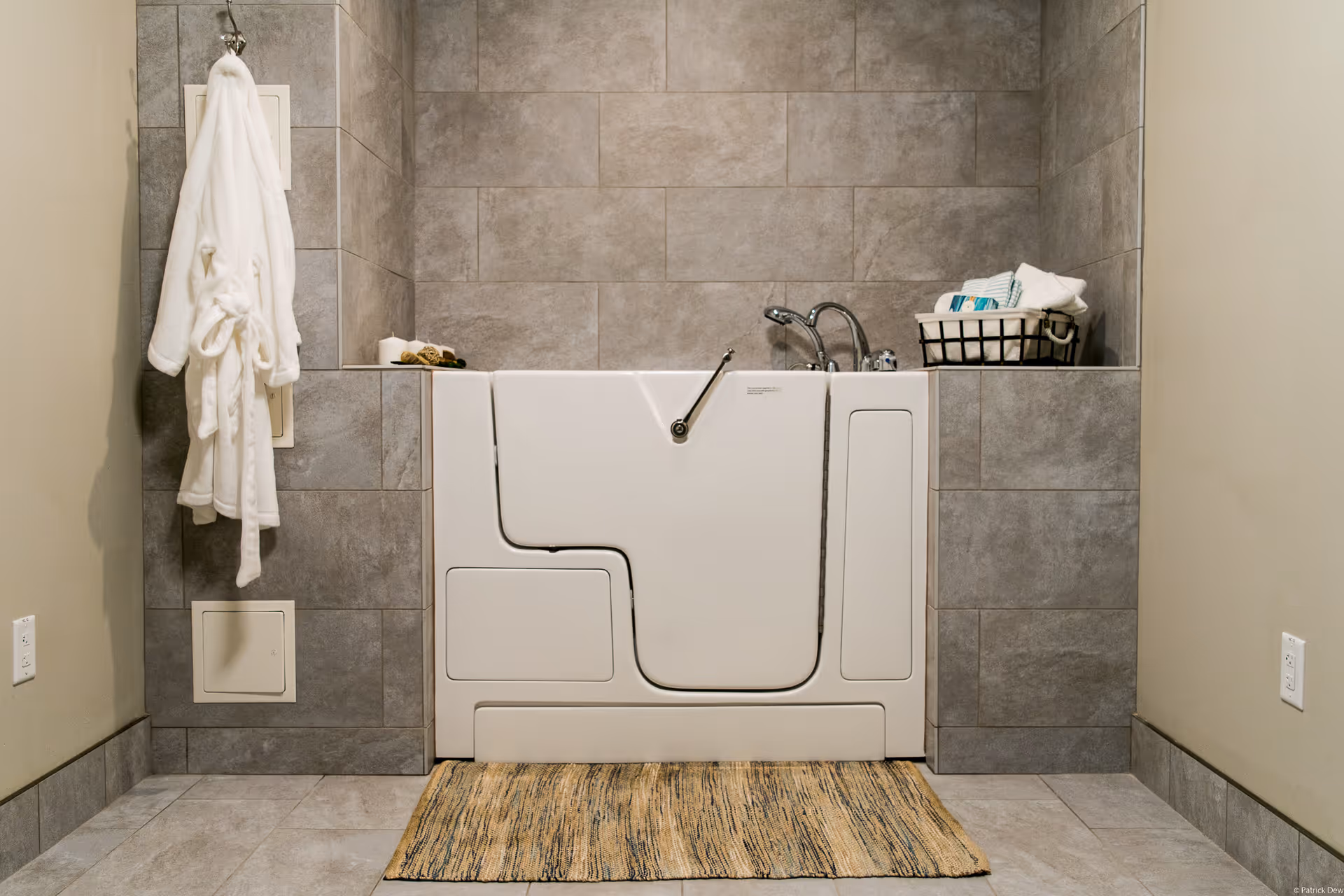 A walk-in bathtub with a closed door, surrounded by gray tiled walls. A white bathrobe hangs on a hook to the left, and a basket with towels and toiletries is placed on the right ledge. A beige and brown striped rug is on the floor in front of the tub.