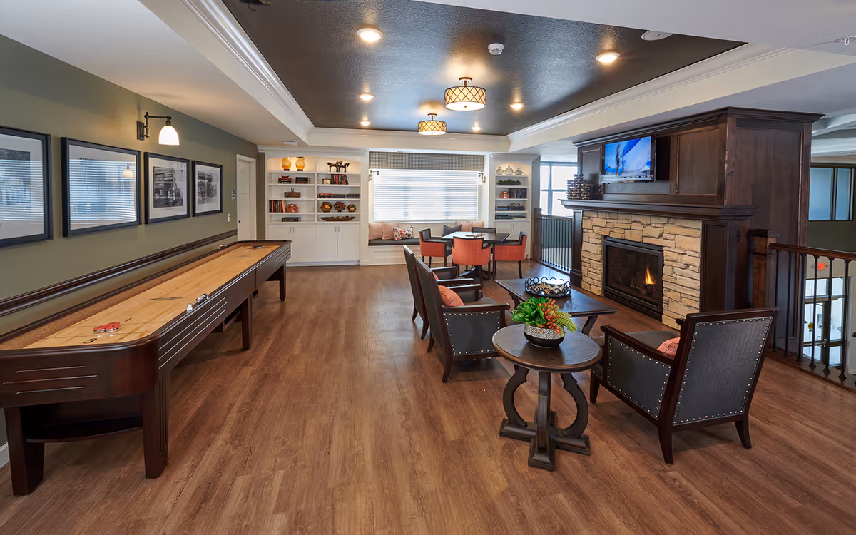 A cozy senior living facility common area with a shuffleboard table on the left, a seating area with armchairs and a coffee table in front of a stone fireplace with a mounted TV above it, and a dining table with chairs near a window with built-in shelves on either side.