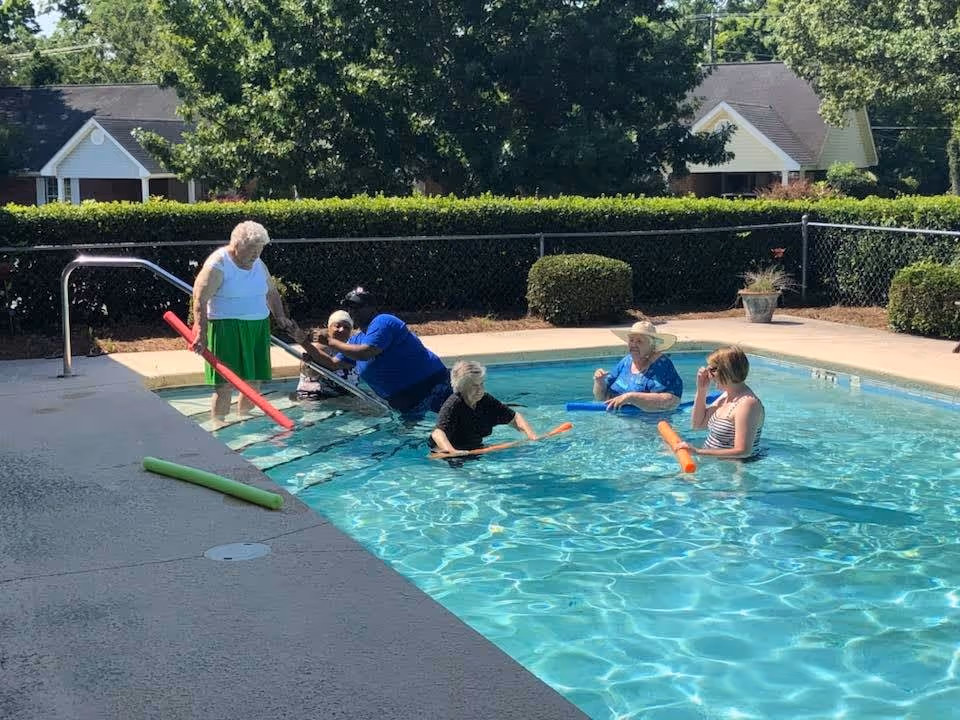 A group of elderly people and a caregiver enjoying time in an outdoor swimming pool. Some are standing or sitting in the water holding colorful pool noodles. The pool is surrounded by a concrete deck, green hedges, and trees with houses visible in the background.