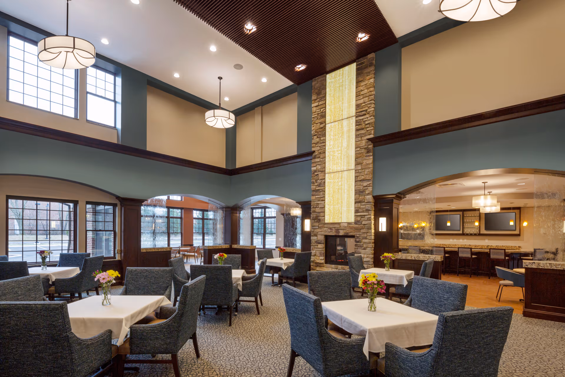 Spacious, well-lit dining and lounge area with tables and upholstered chairs, a tall stone fireplace feature and high ceilings with pendant lights.