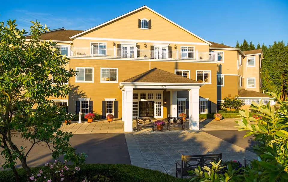 Exterior view of Canterbury Park Independent Living Community building with a covered entrance, multiple windows, and a well-maintained garden with trees and flowers in the foreground under a clear blue sky.