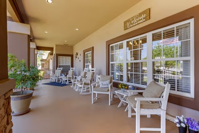 A spacious covered porch area with several white rocking chairs and small tables arranged along the wall. There are potted plants and flowers placed around, and a wooden sign above the window reads 'Welcome to our porch'. The porch overlooks a green outdoor area visible through the windows.