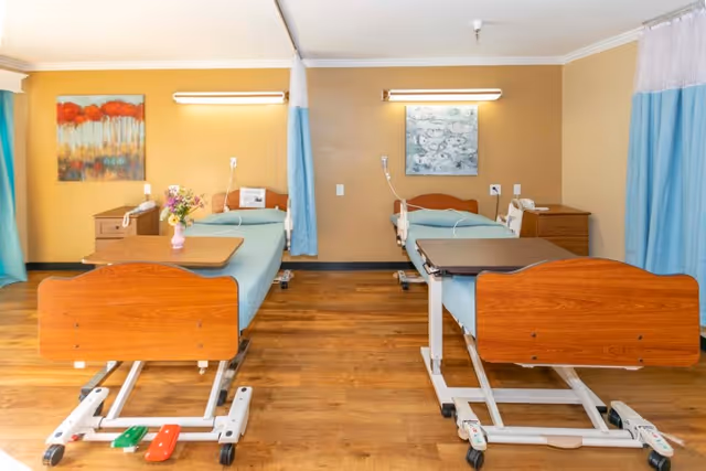 Two hospital-style beds with wooden headboards in a shared patient room separated by curtains, each with bedside tables, overhead lights, and a wood floor.